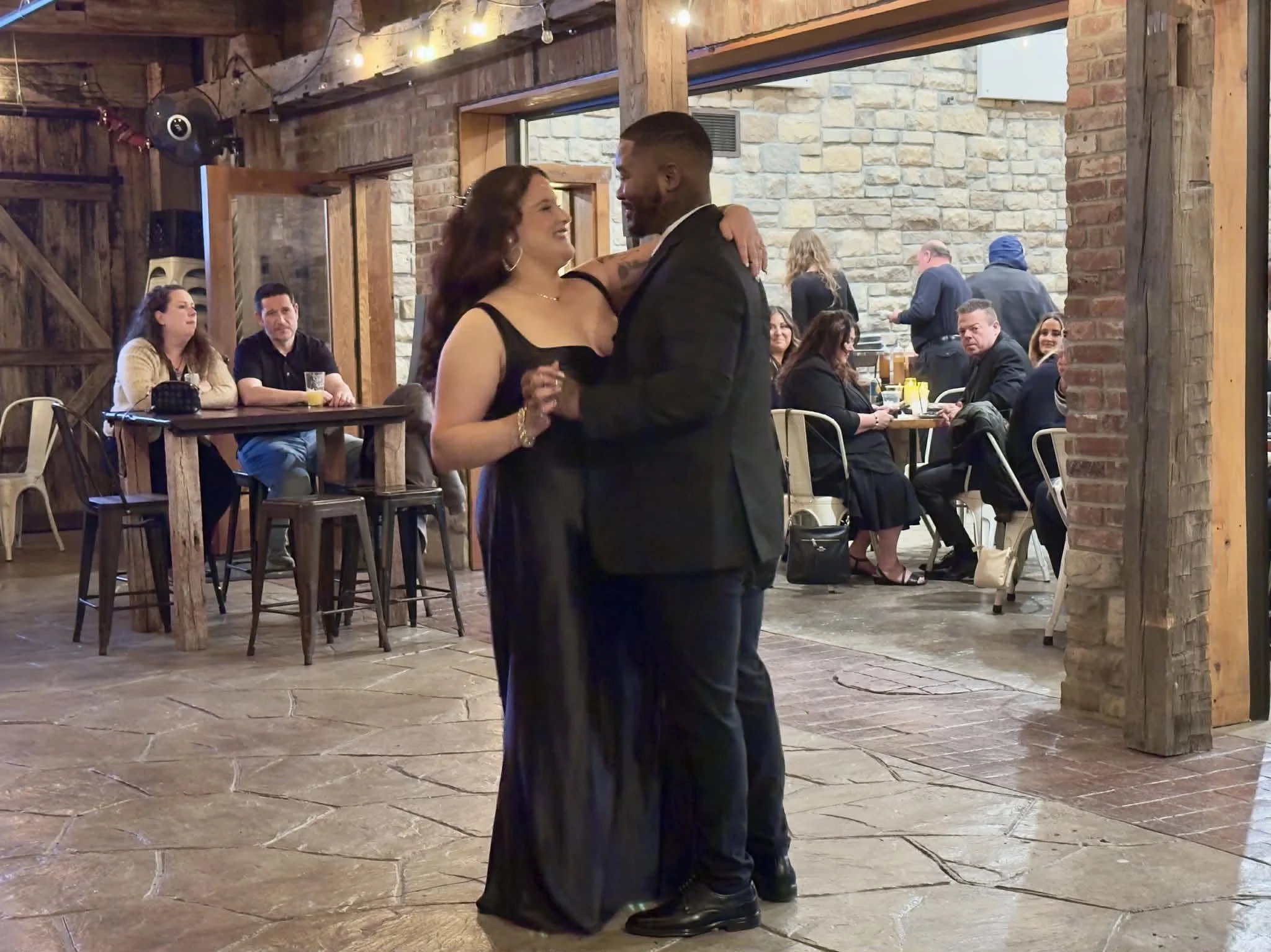 A couple dancing together in a rustic, wood and brick decorated venue with several seated guests watching.