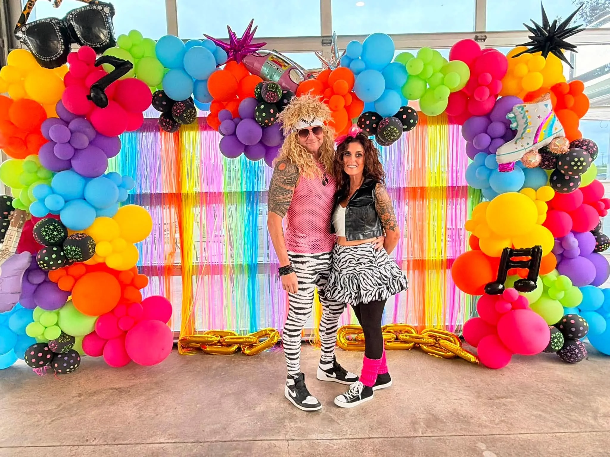 Two people dressed in zebra print clothing standing in front of a colorful balloon backdrop, decorated with rainbow streamers and rainbow-colored large balloons, with number 2 and 1 balloons on the floor.