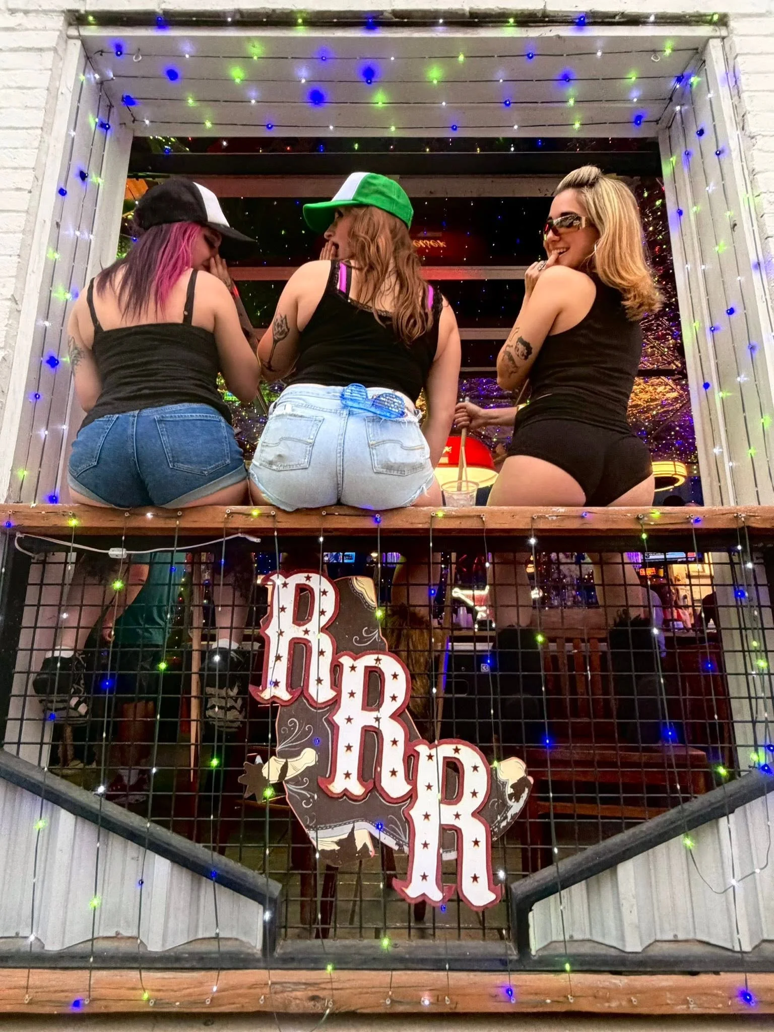 Three women sitting on a porch with colorful Christmas lights and a "Designed by Dad" sign hanging below.