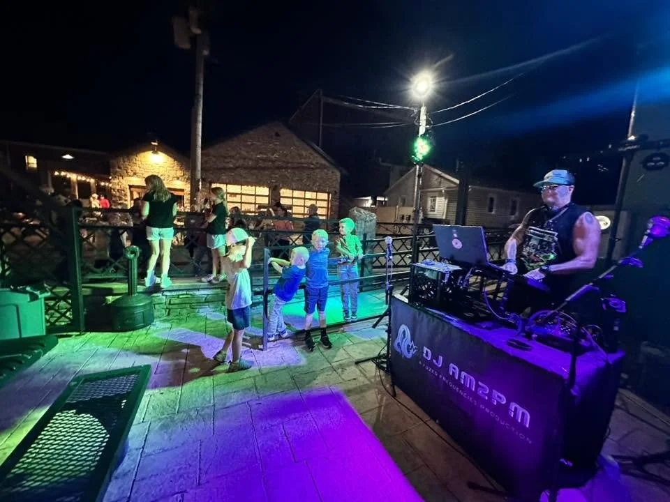 Nighttime outdoor dance party with children dancing on a wooden deck, a DJ playing music, and colorful lighting.