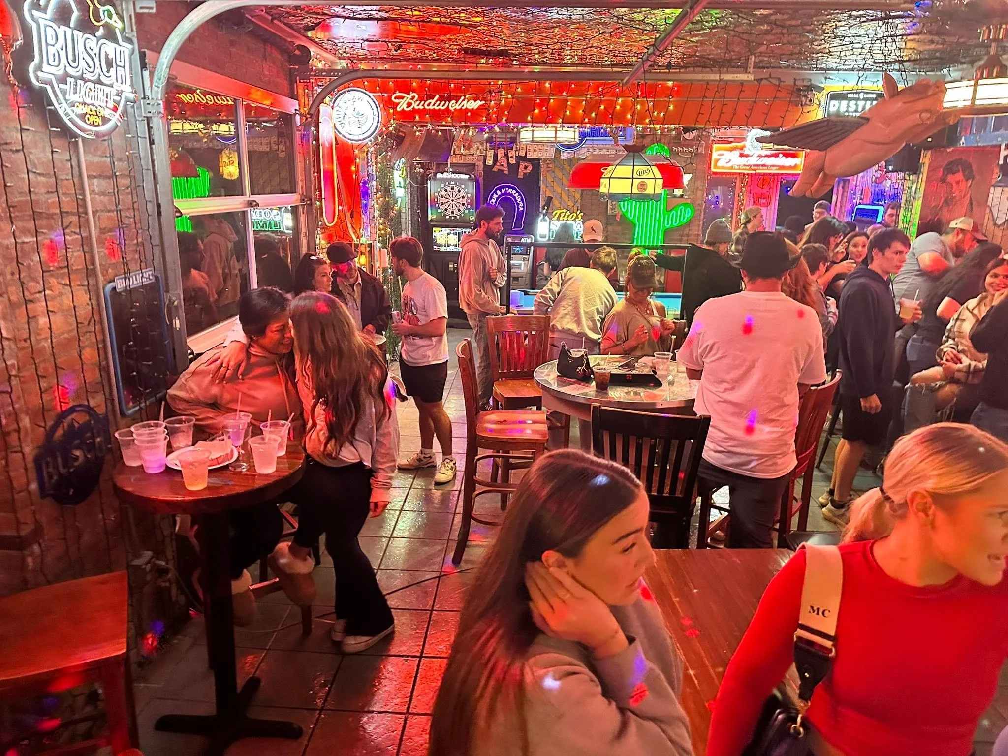 A crowded bar or pub decorated with numerous neon signs, string lights, and neon-colored decorations, including a large red cactus with a sombrero. People are socializing, standing, and sitting at tables with drinks. The atmosphere appears lively and