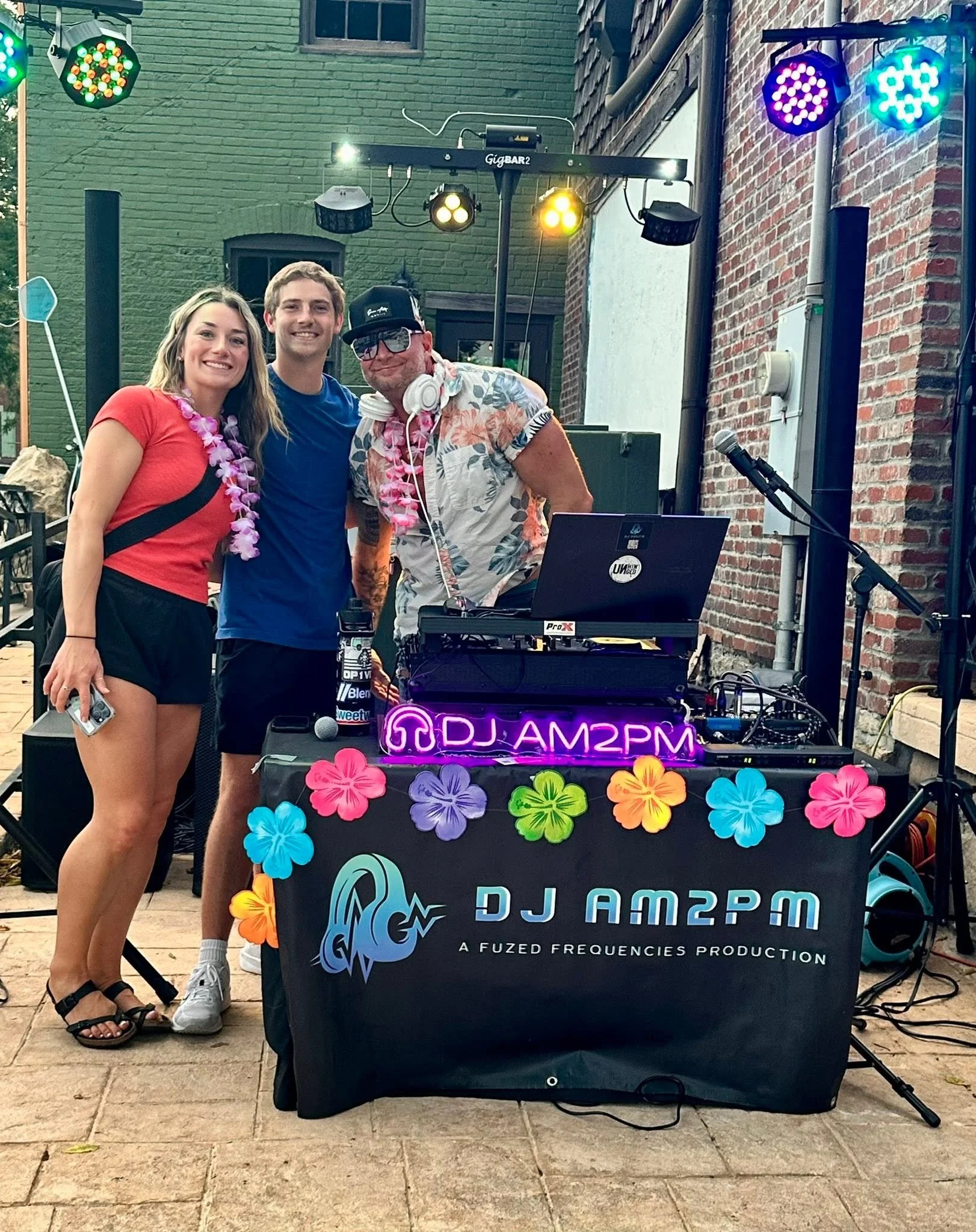 Three people standing behind a DJ booth with colorful flower decorations and neon sign reading 'DJ AM2PM'. The woman on the left is wearing a red shirt, black shorts, sandals, and a pink lei. The man in the middle is dressed in a blue shirt, white sh