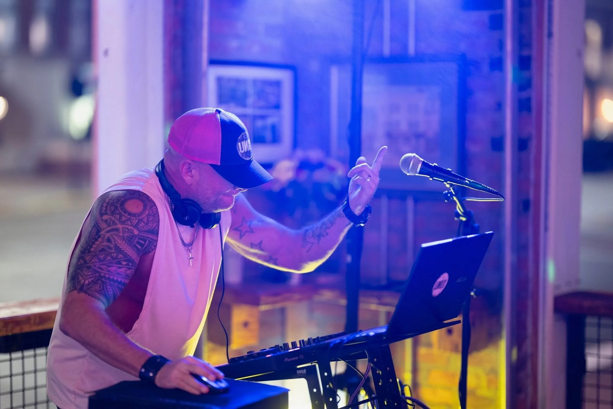 Man with tattoos on his arm, wearing a pink tank top, baseball cap, and headphones around his neck, pointing his finger while working with DJ equipment at a club with colorful lighting.