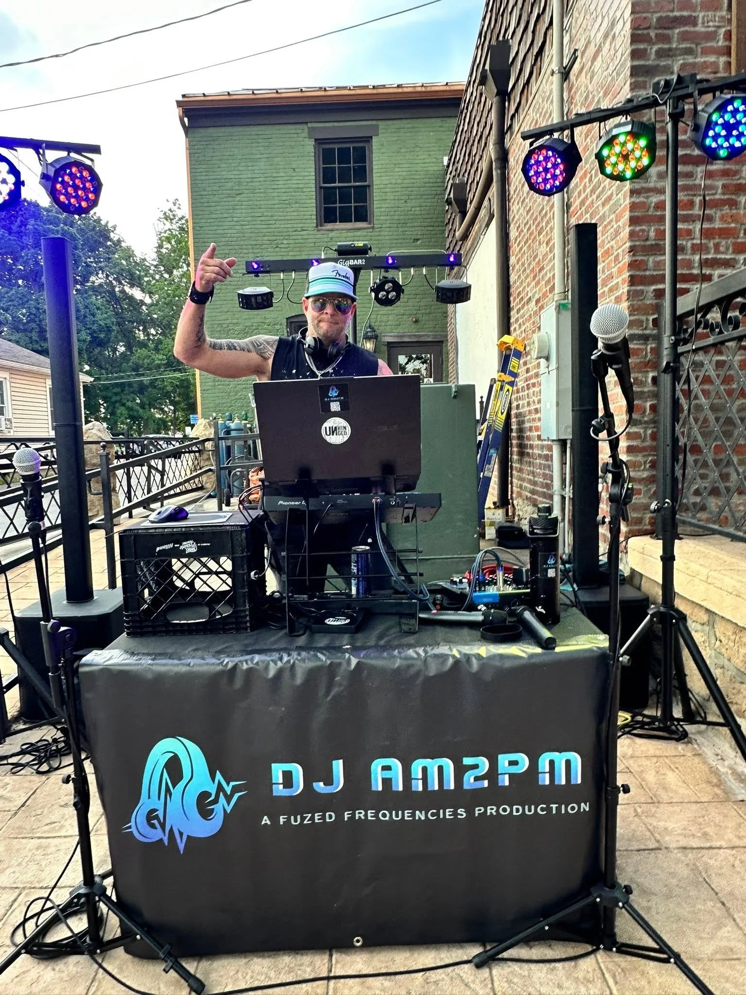 A DJ standing behind a DJ booth in an outdoor setting, with colorful stage lights overhead and brick buildings in the background. The DJ is wearing sunglasses, a cap, and has tattoos on his arms.