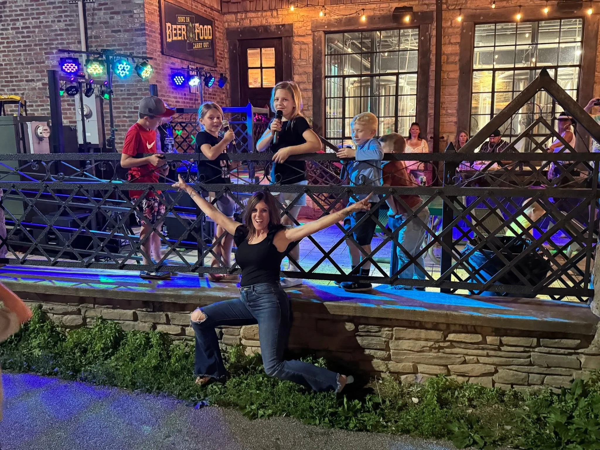 Children performing karaoke on a stage with colorful lights in a brick-walled venue, with adults and a woman posing in front.