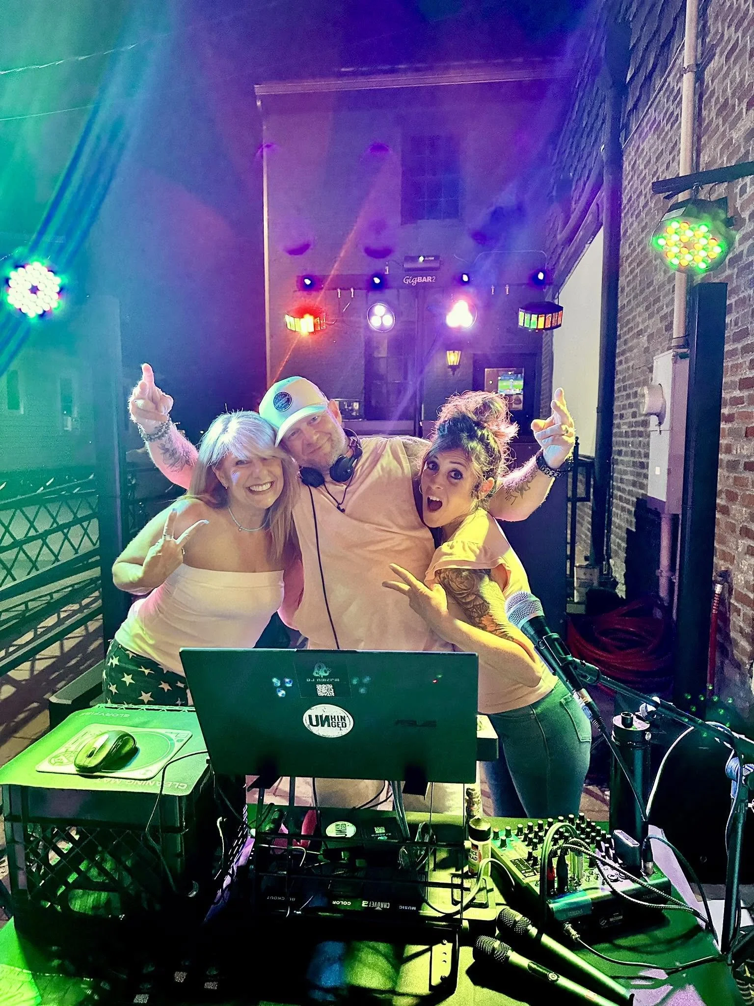 Three people at a DJ booth on a stage with colorful lighting, DJ equipment, and a microphone, dancing and posing together.