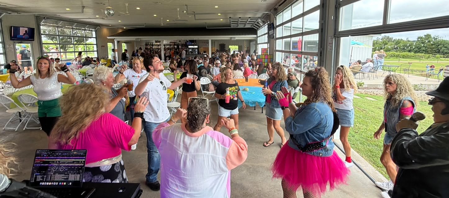 People dancing and celebrating indoors at a party, with some outside visible through the large windows.