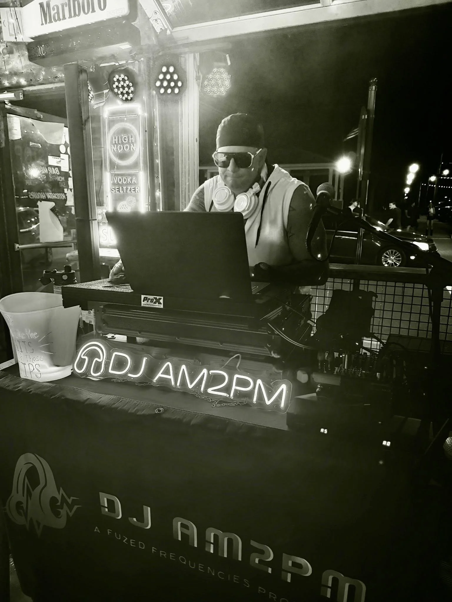 A DJ wearing sunglasses, a hoodie, and headphones around their neck working at a DJ booth in an outdoor setting at night. Neon signs and people are visible in the background.