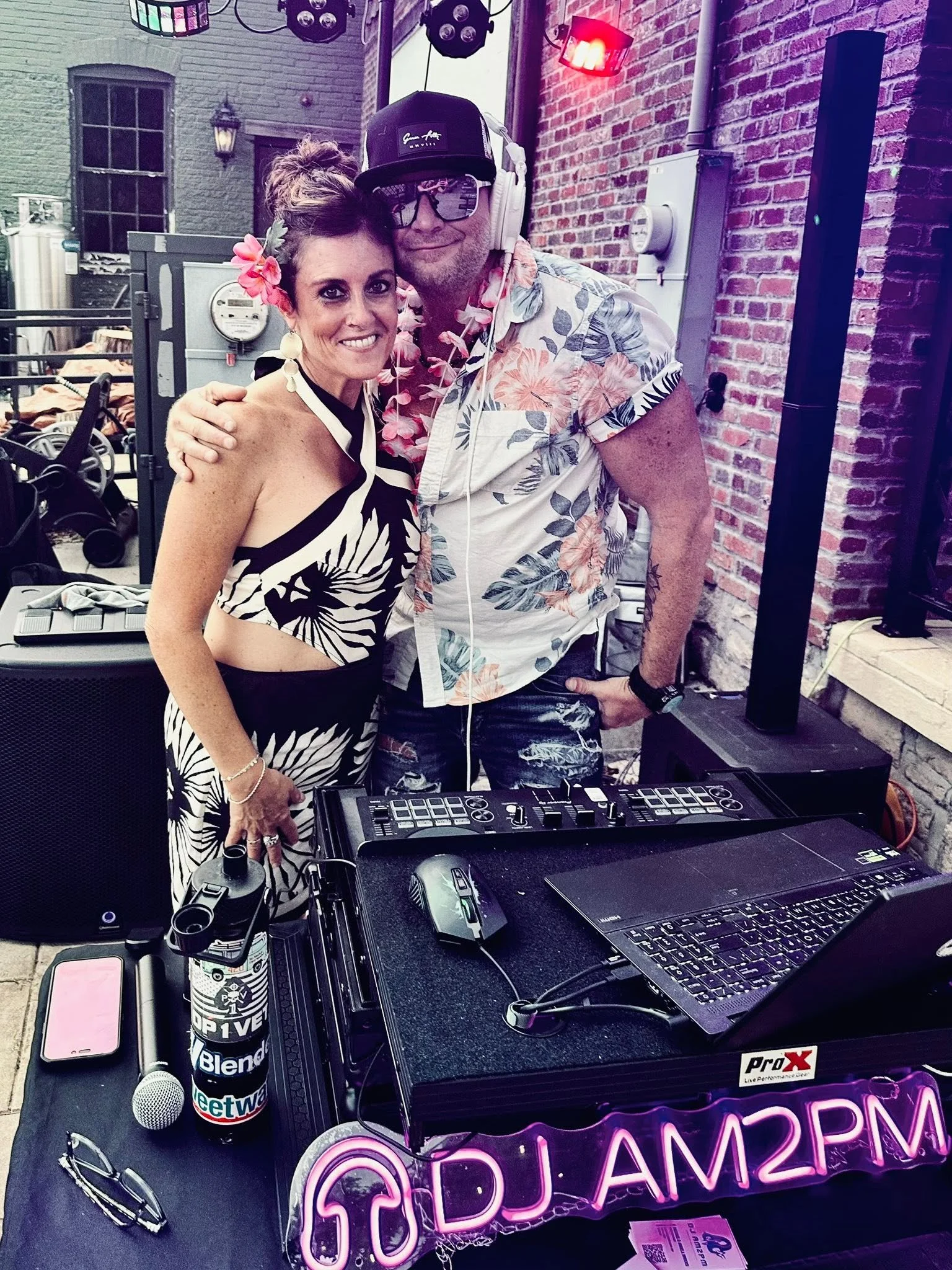 Two people smiling and posing together at a DJ setup in an outdoor urban setting with brick walls. The woman has flowers in her hair and is wearing earrings, while the man has headphones and sunglasses, and is wearing a floral shirt. DJ equipment and