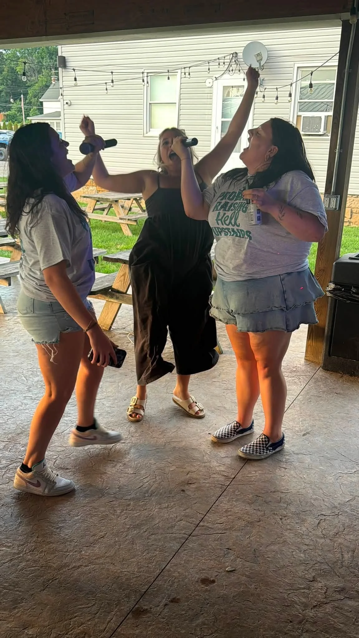 Three women singing and dancing together on a covered porch with picnic tables and string lights in the background.