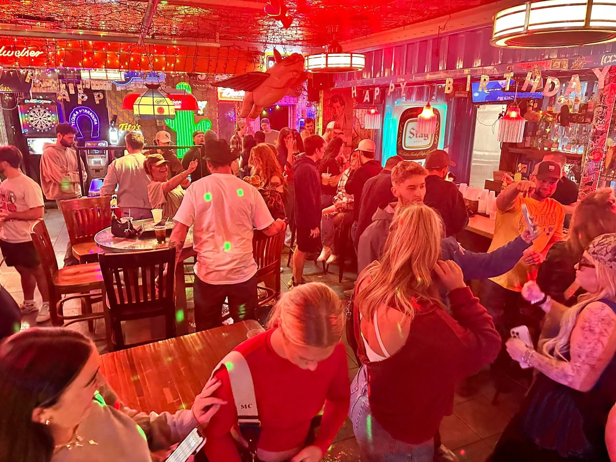 Crowd of people at a lively bar or nightclub with neon signs, colorful lights, and a 