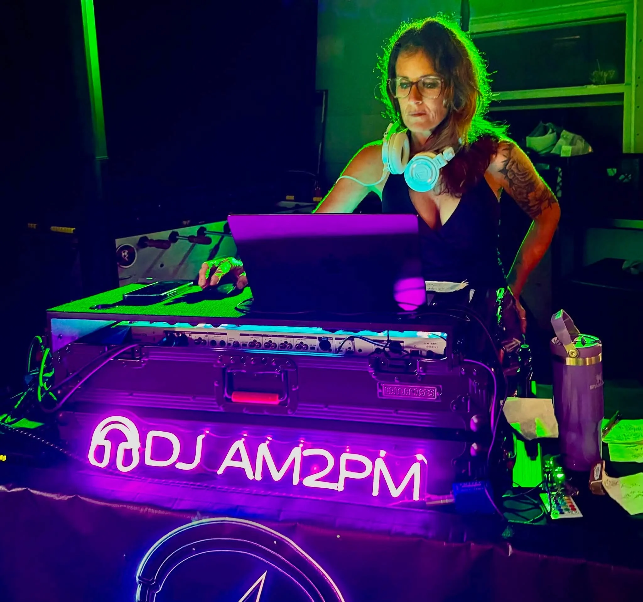 A woman with glasses and tattooed arm working at a DJ booth illuminated by colorful neon lights with a sign displaying Greek letters.