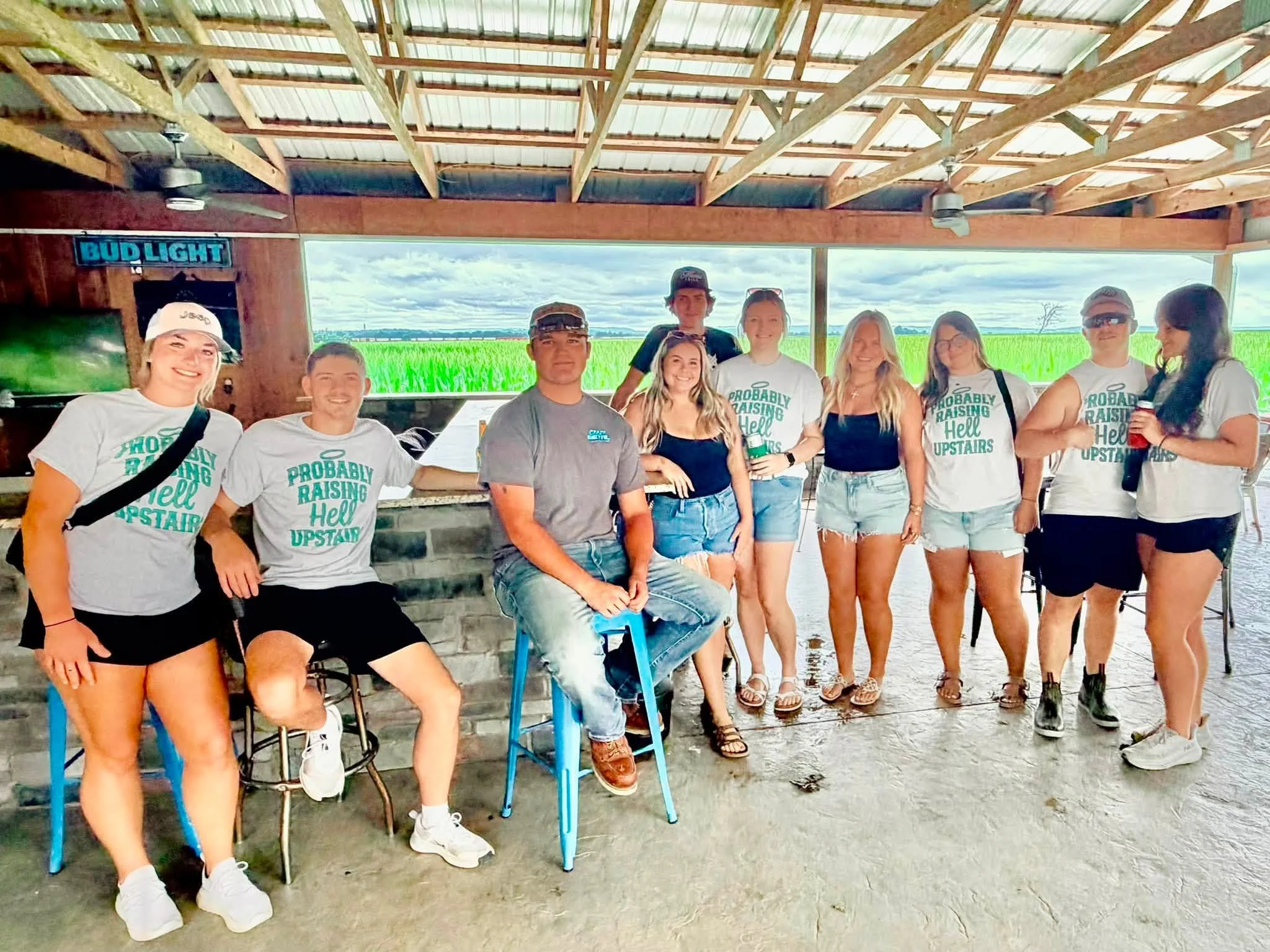 A group of people standing together under a covered outdoor area with a view of a field and cloudy sky in the background. Some members are wearing white T-shirts with the phrase 'Probably Raising Hell Upstairs' printed on them.