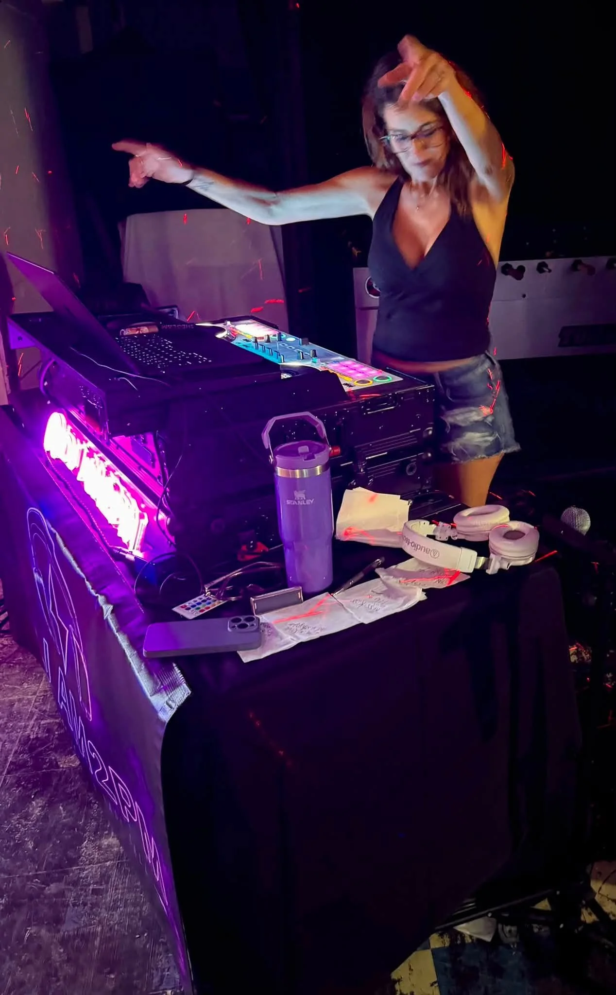 A woman DJ wearing glasses and a black tank top, standing behind a DJ booth with colorful lights, music equipment, and headphones, in a dark setting, possibly at a club or party.