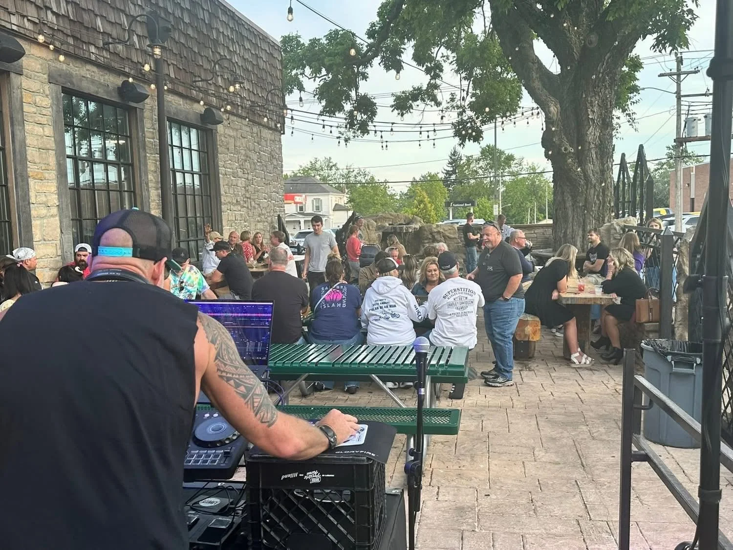 Outdoor gathering at a restaurant with people seated at tables, a DJ playing music in the foreground, and string lights hanging above under a large tree.