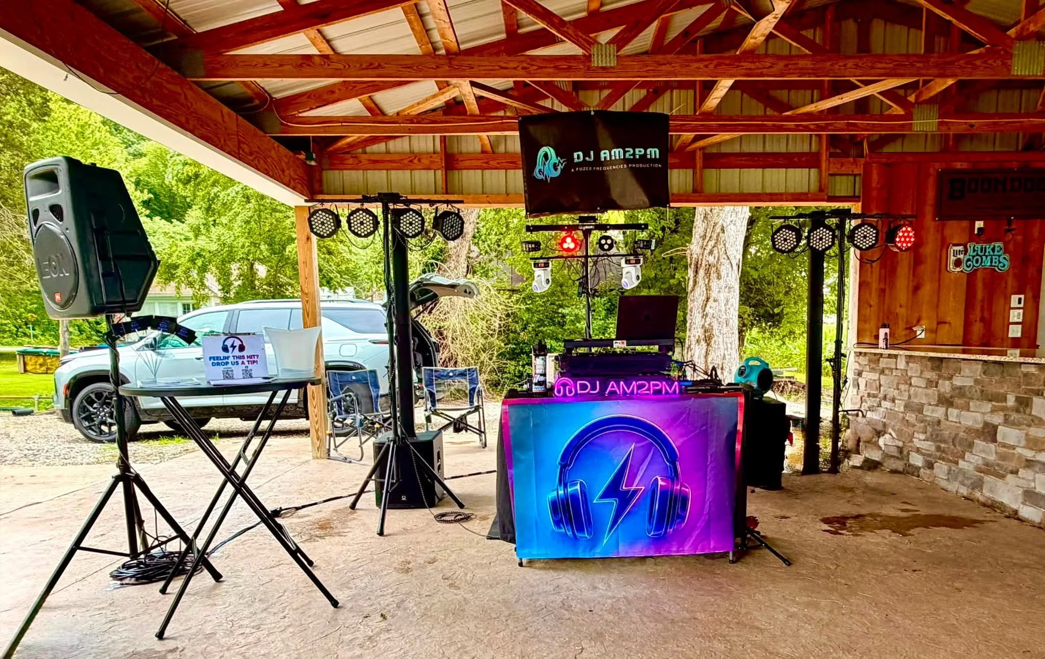 An outdoor stage with DJ equipment, speakers, lighting, and a neon sign at a venue with a wooden roof and surrounded by trees and parked cars.