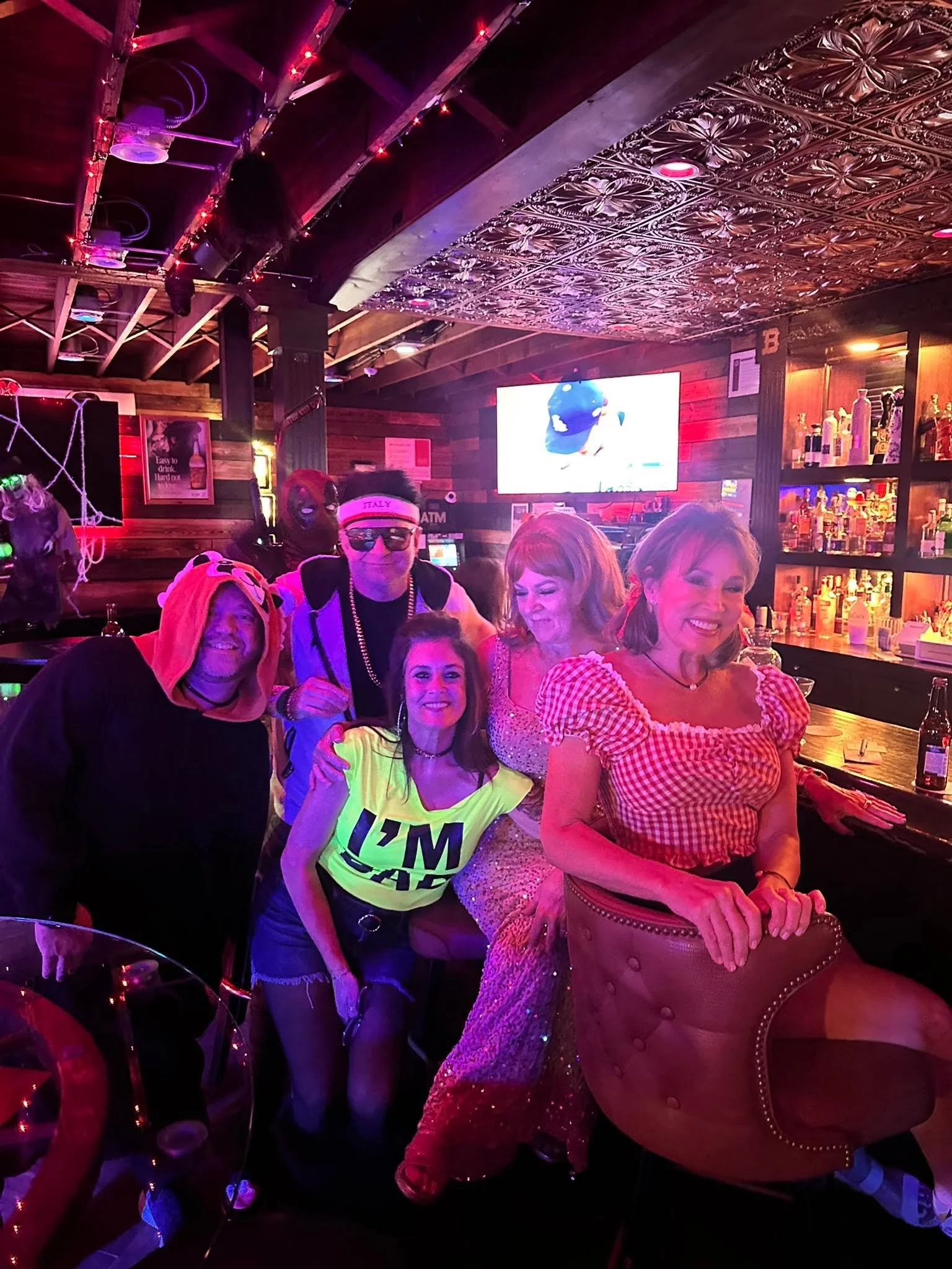 Group of people in a bar or nightclub with a festive atmosphere, colorful lighting, and a bar shelf with bottles in the background.