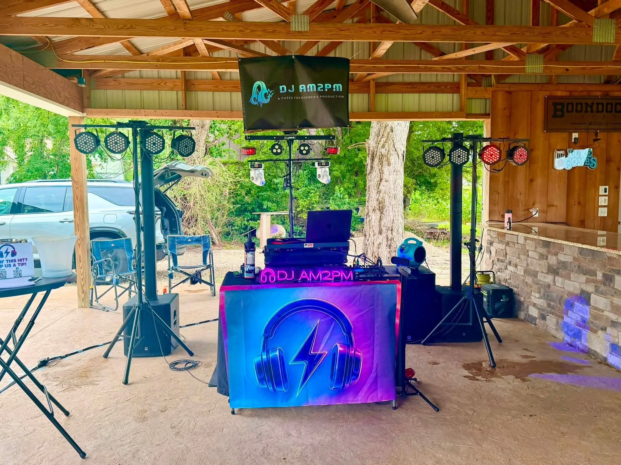 Outdoor music setup with DJ equipment, stage lighting, and a large screen displaying DJ AM2PM, under a wooden pavilion with a corrugated metal roof, surrounded by green trees and parked cars.