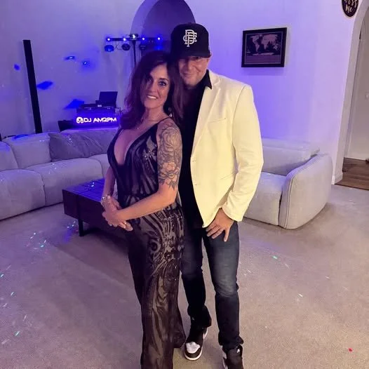 A woman in a sheer black dress and a man in a white blazer and black cap standing together in a living room with a white couch and a party setup