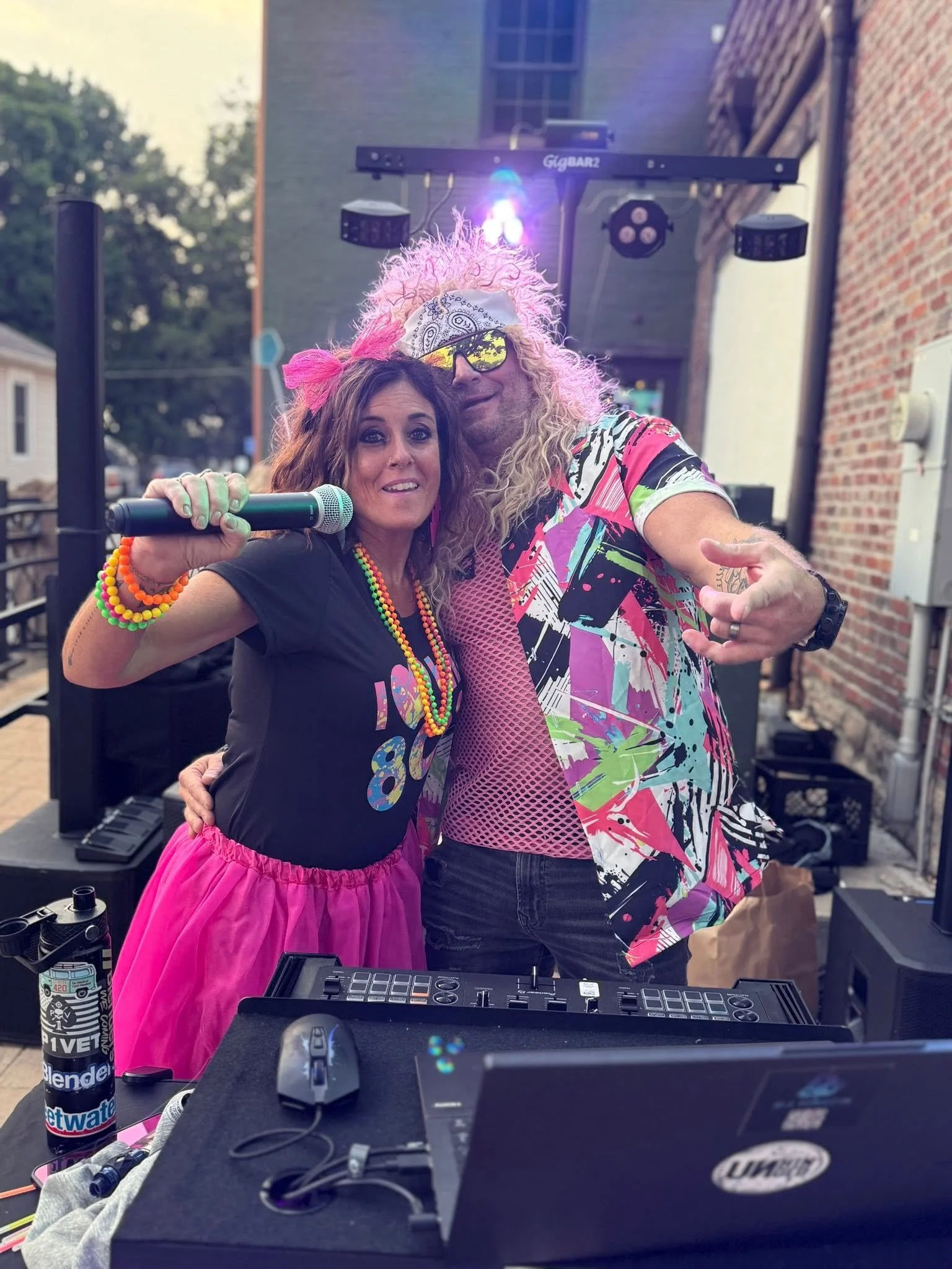 Two people at an outdoor event taking a selfie. The woman is wearing a black t-shirt and pink skirt, holding a microphone, with colorful beaded necklaces and bracelets. The man is wearing a vibrant, patterned shirt, yellow mirrored sunglasses, and a 