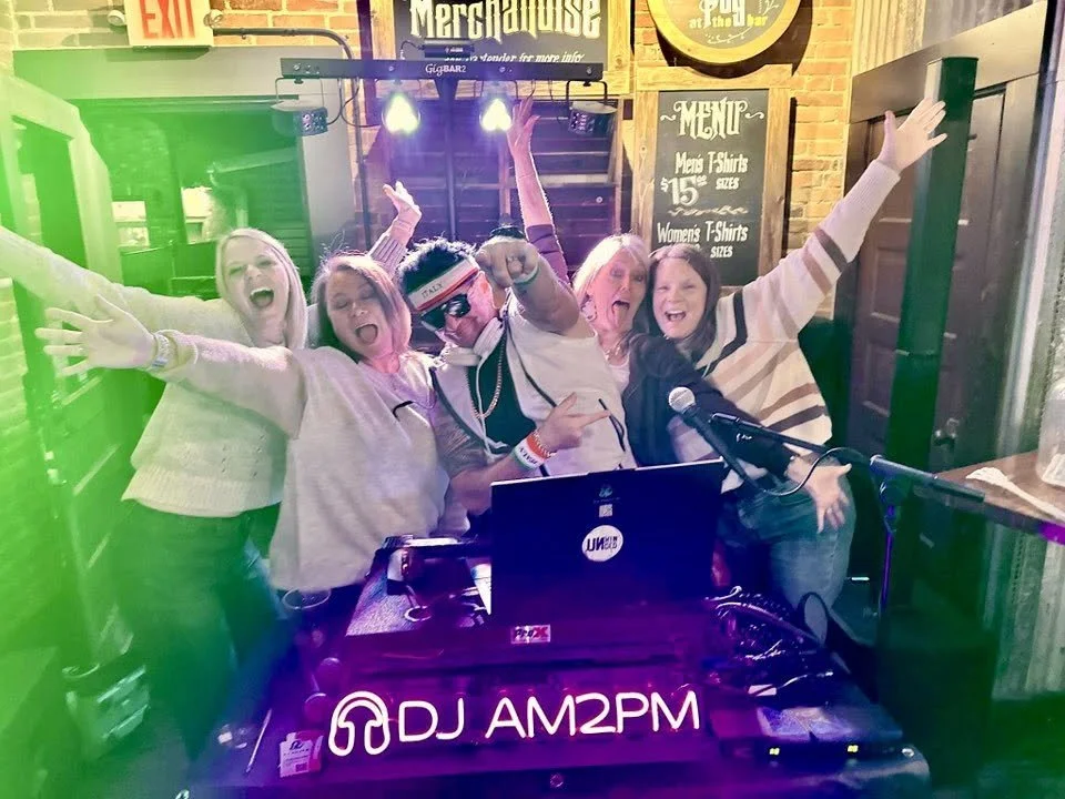 Group of five people celebrating at a DJ booth in a bar or restaurant, with one person wearing sunglasses and a visor, and others smiling and raising their hands.