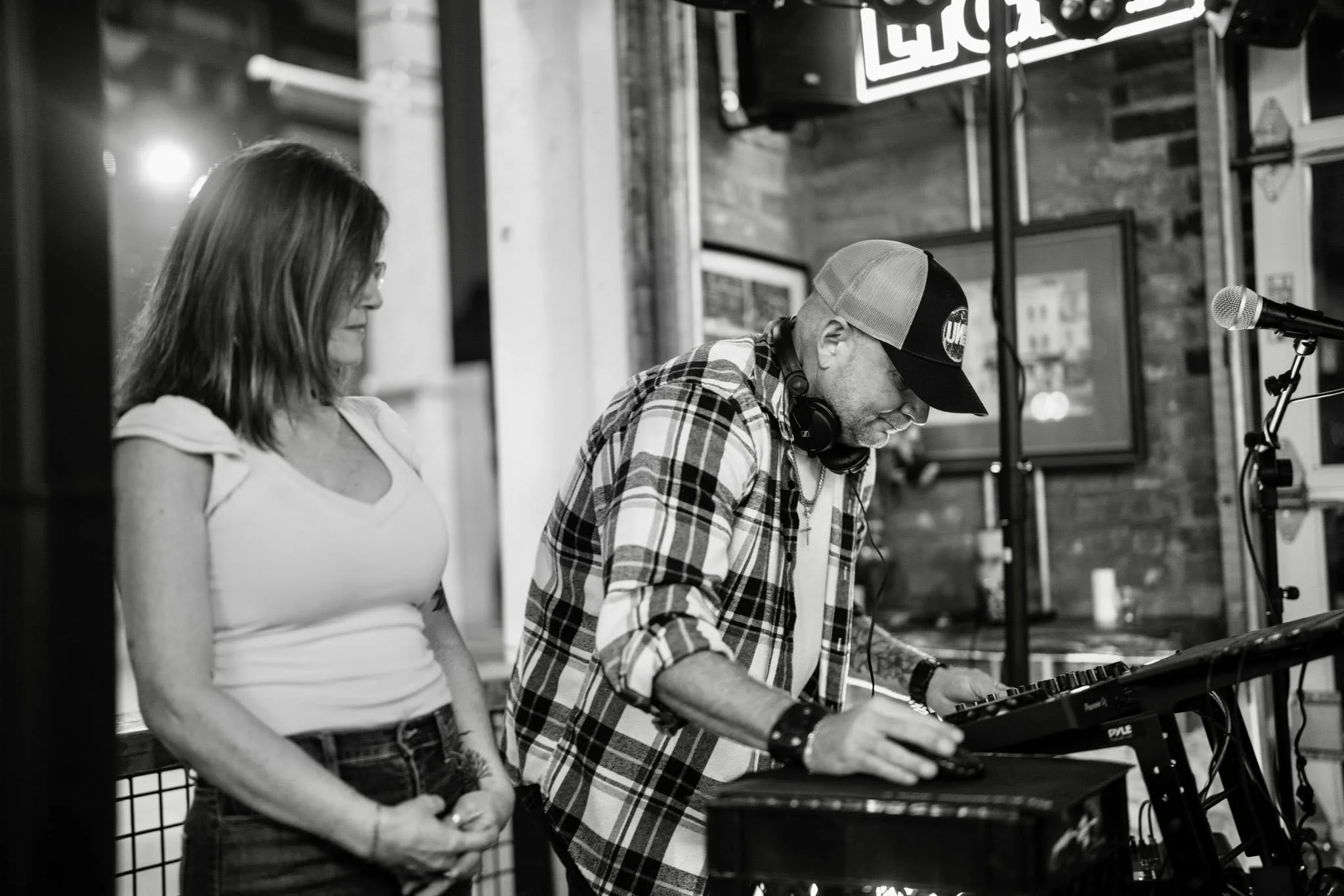 A woman and a man with a baseball cap and headphones, both focused on a music controller, in a casual indoor setting.