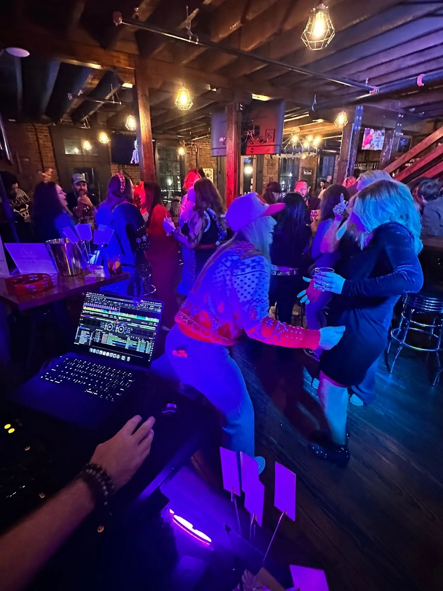 People enjoying a night out at a bar or nightclub with a DJ playing music, exposed brick walls, and warm ambient lighting.