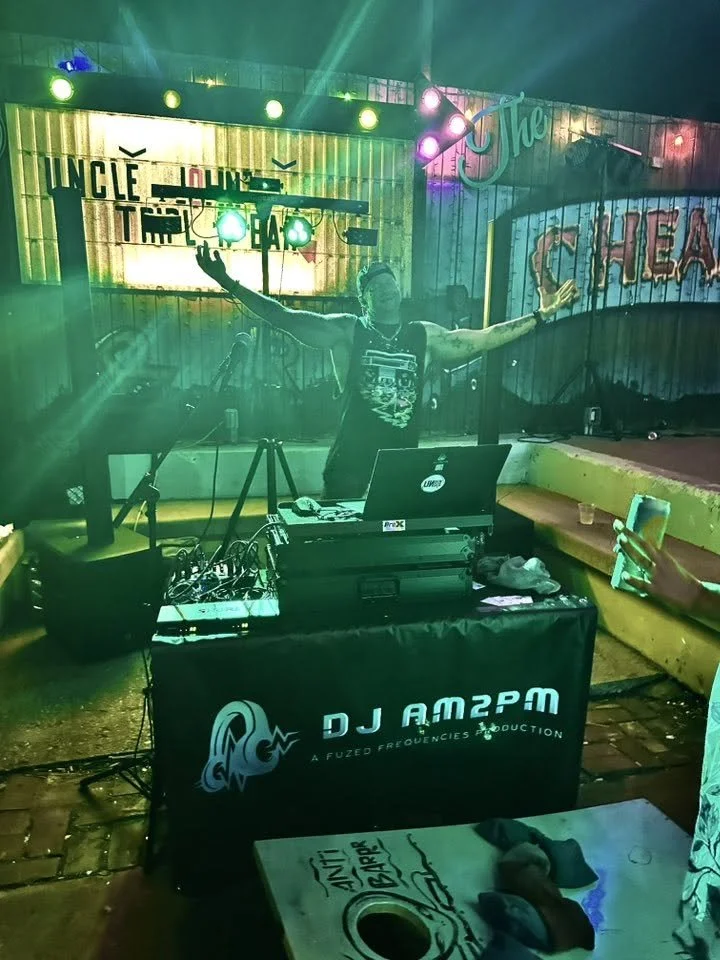 DJ performing at a nightclub or bar with colorful lighting. The DJ is standing behind a table with DJ equipment, wearing a sleeveless shirt and headphones. Neon signs and a stage backdrop are visible in the background.