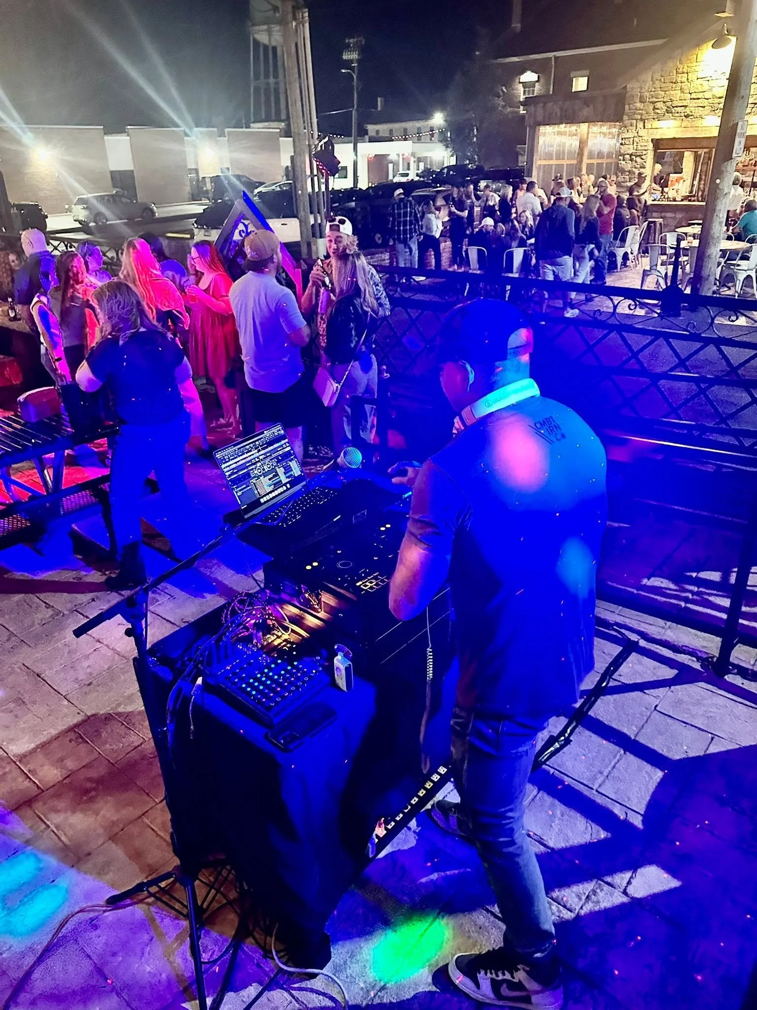 A DJ performing at an outdoor night party with a crowd of people dancing and socializing. The setting includes colorful lights, a table with DJ equipment, and a street with parked cars.