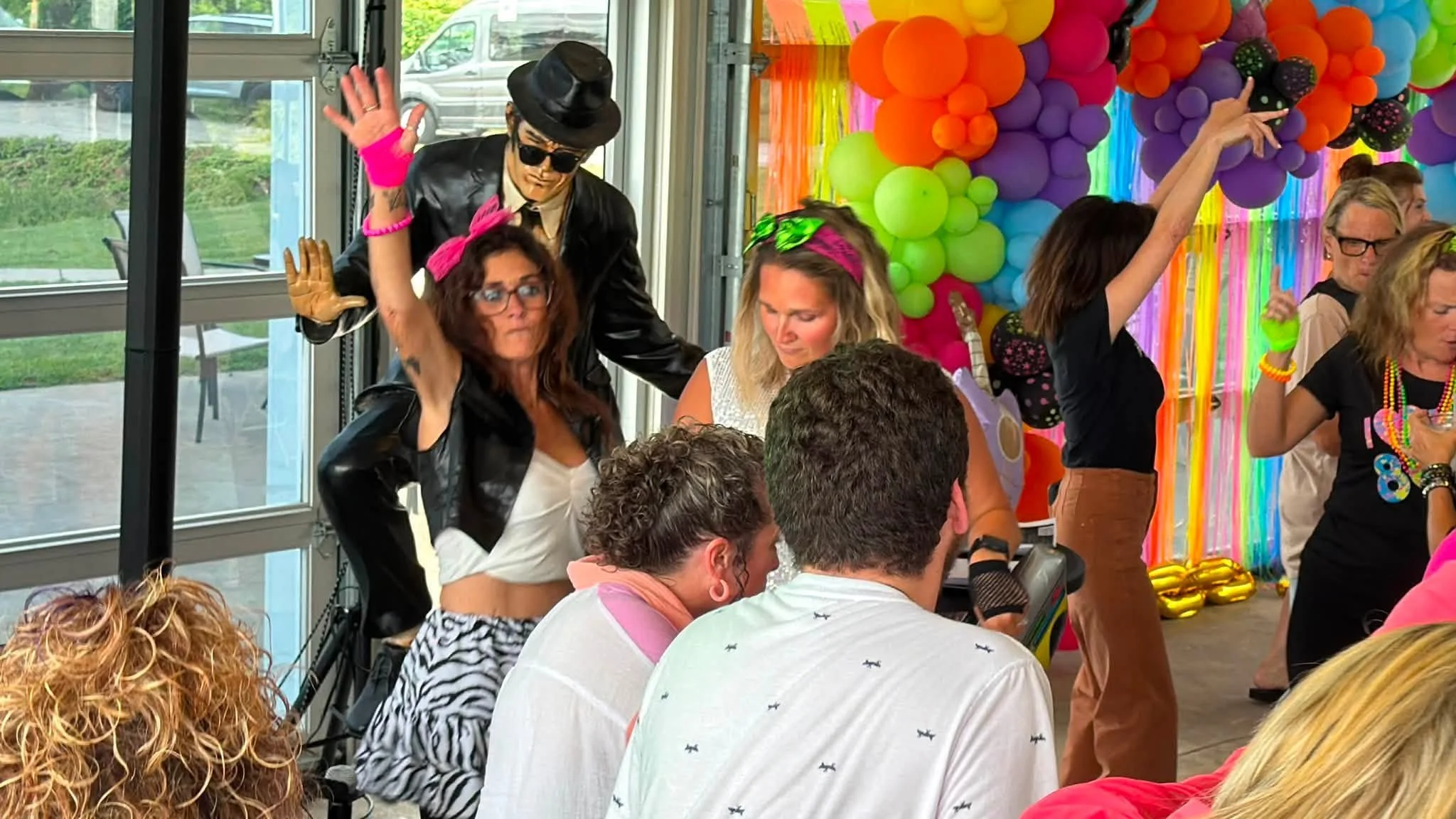 People gathered indoors at a colorful party with balloon decorations, some wearing vibrant accessories, and a person dressed as Elvis Presley.