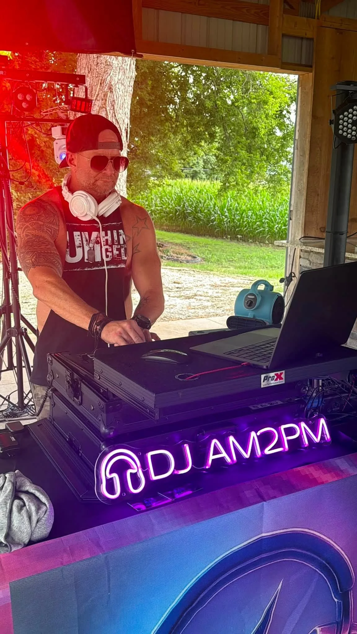 A DJ wearing sunglasses, a backwards cap, and headphones around his neck, standing behind a DJ setup with a laptop and equipment, with a neon sign displaying the word 'DREAM' in purple and pink lights, outdoors with trees and cornfield in the backgro