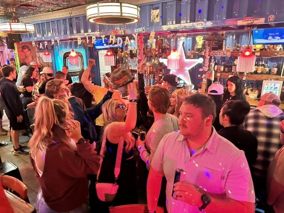 People gathering in a lively bar with colorful neon lights, birthday decorations, and a crowded bar counter in the background.
