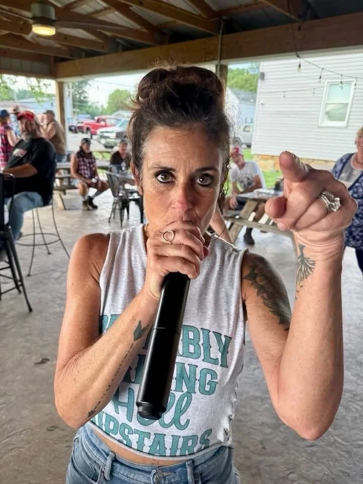 A woman with dark hair in a bun, wearing a sleeveless shirt, holds a microphone close to her mouth and points directly at the camera, with a group of people sitting at tables in the background at an outdoor gathering under a pavilion.