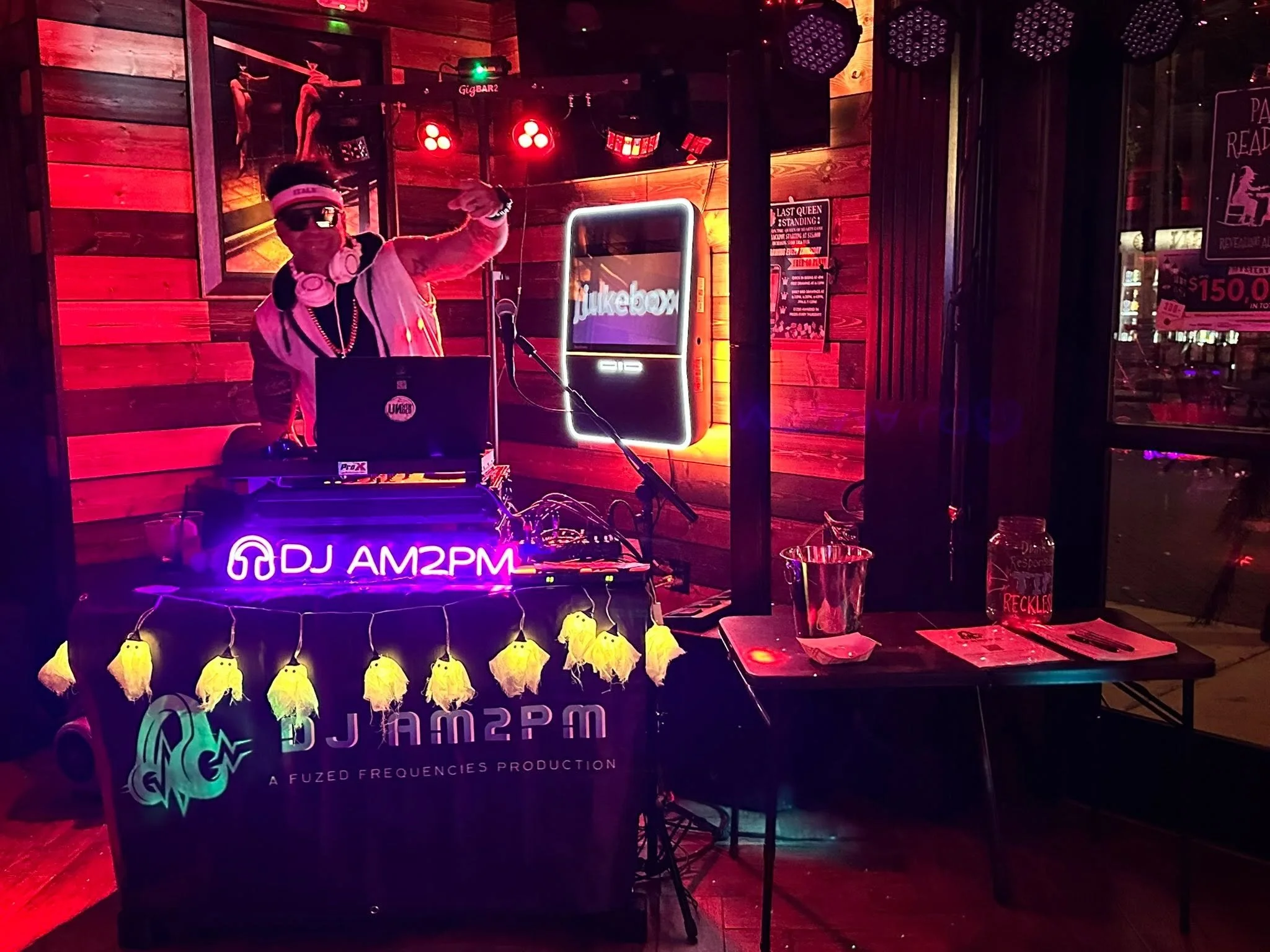 A DJ performing at a nightclub with red lighting, wooden wall decor, a neon sign reading 'jukebox,' and Halloween decorations including yellow ghost hanging lights. The DJ is wearing sunglasses and headphones, with a laptop and sound equipment in fro