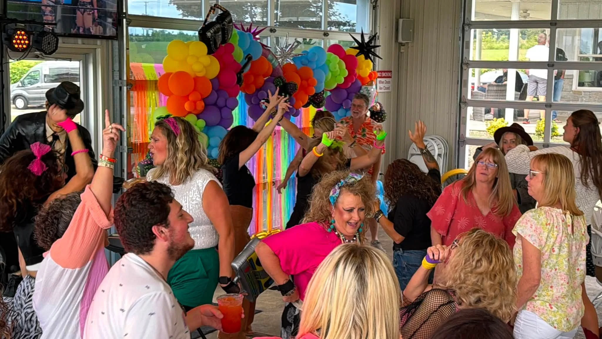 A lively party scene with people dancing and celebrating in a decorated indoor space featuring colorful balloon arrangements and rainbow-themed decorations.