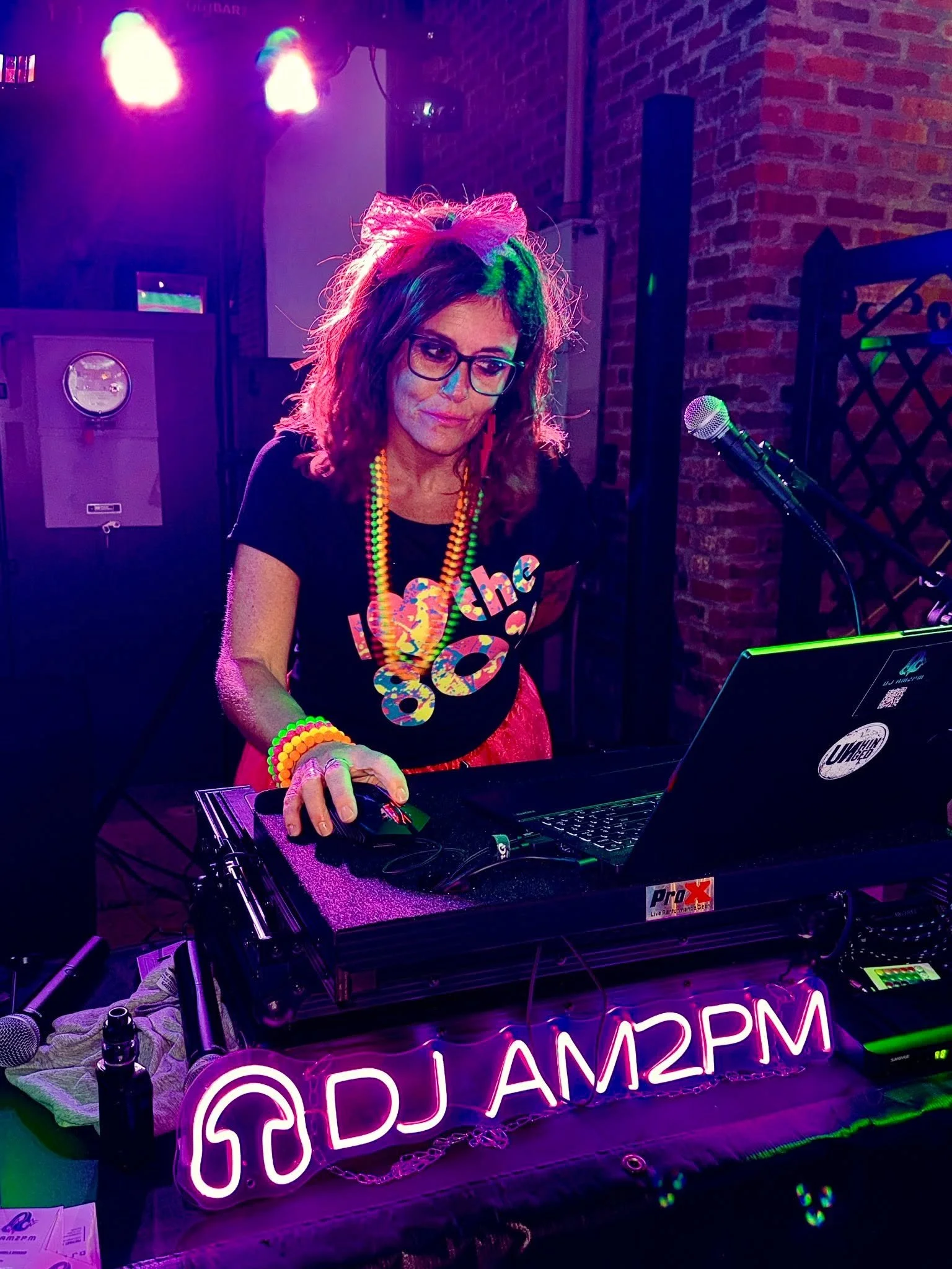 A woman wearing glasses, colorful beads, and a black t-shirt with a rainbow-colored heart, working at a DJ booth with a neon sign that says 'DJ AM2PM' in pink and purple lights, surrounded by equipment and a brick wall.