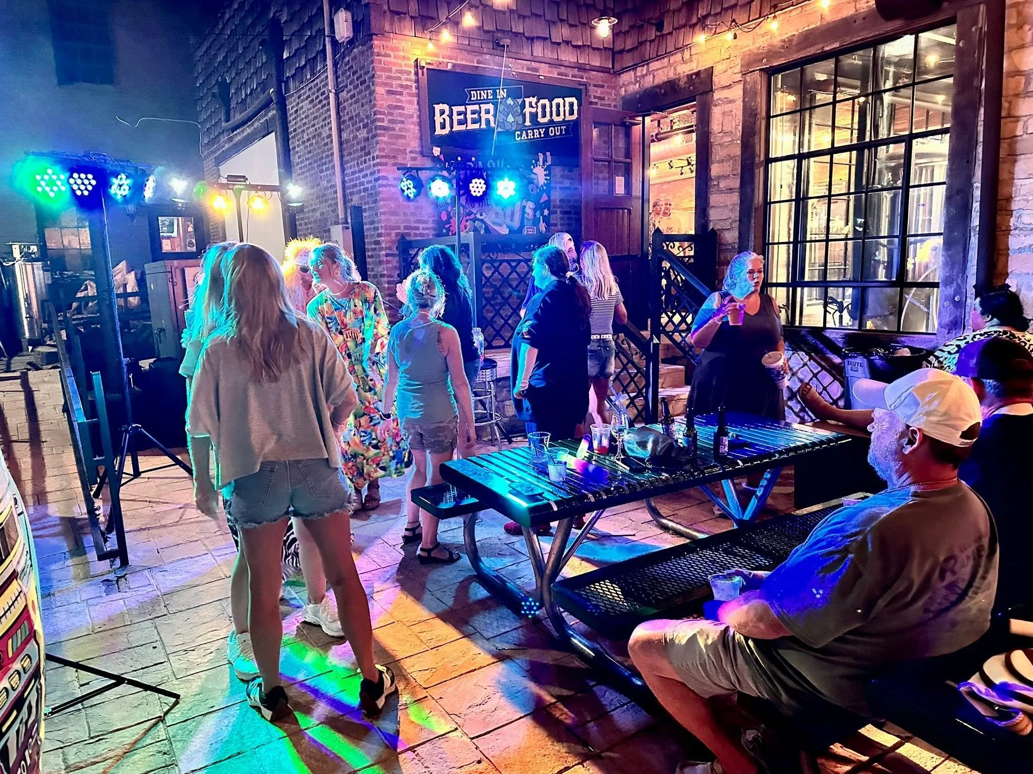 A group of people socializing outside a brick building with large windows, illuminated by colorful lights at night, with a sign that reads 'Dine In, Beer, Food, Carry Out.'