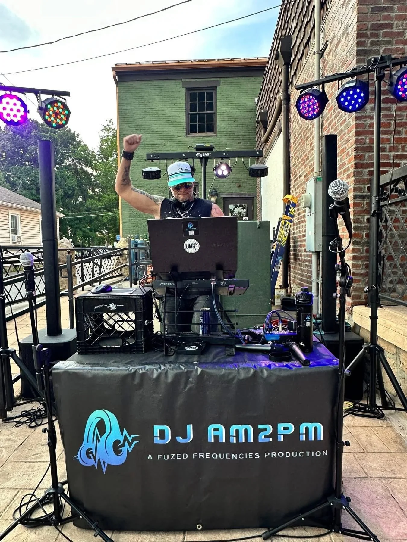 A DJ wearing sunglasses and a baseball cap is performing outdoors on a stage with DJ equipment, including a laptop, mixer, and microphones set up on a table with a DJAM2PM logo. The DJ is raising one fist in the air, and the stage has colorful lighti