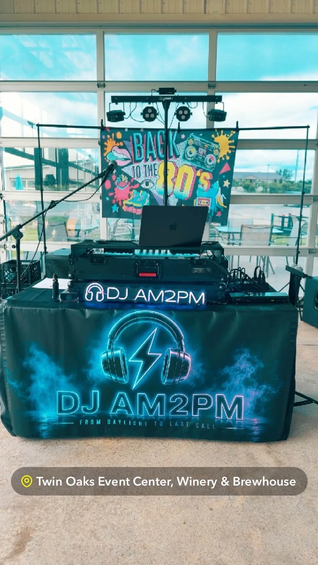 A DJ setup at Twin Oaks Event Center with a banner that reads 'Back to the 90s' in colorful text, featuring retro graphics including a cassette tape and boombox, along with a neon sign displaying 'DJ AM2PM' and a lightning bolt and headphones logo, i