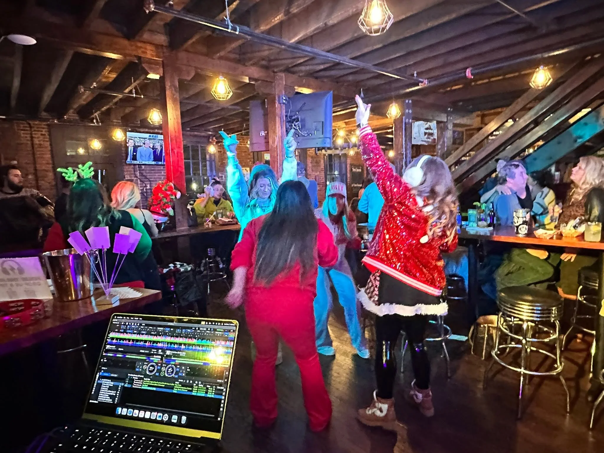 People dancing and celebrating in a rustic bar or club with wooden ceiling beams and warm lighting, including a DJ with a laptop in the foreground.
