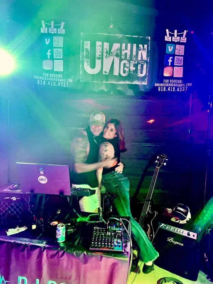 Two people smiling and hugging in front of a DJ setup with a sign that says 'Unhinged' in a colorful, nightclub-like atmosphere.