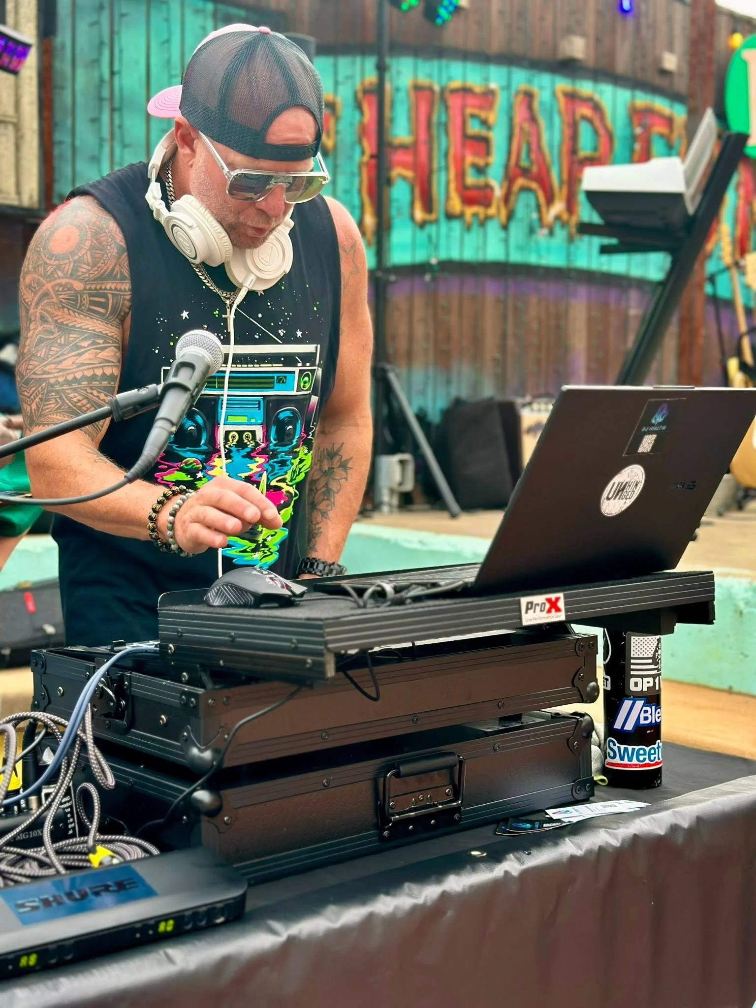 A man with tattoos, wearing a black sleeveless shirt with a colorful boombox graphic design, sunglasses, a cap backwards, and headphones around his neck, standing behind DJ equipment with a laptop, microphone, and cables, in front of a colorful, illu