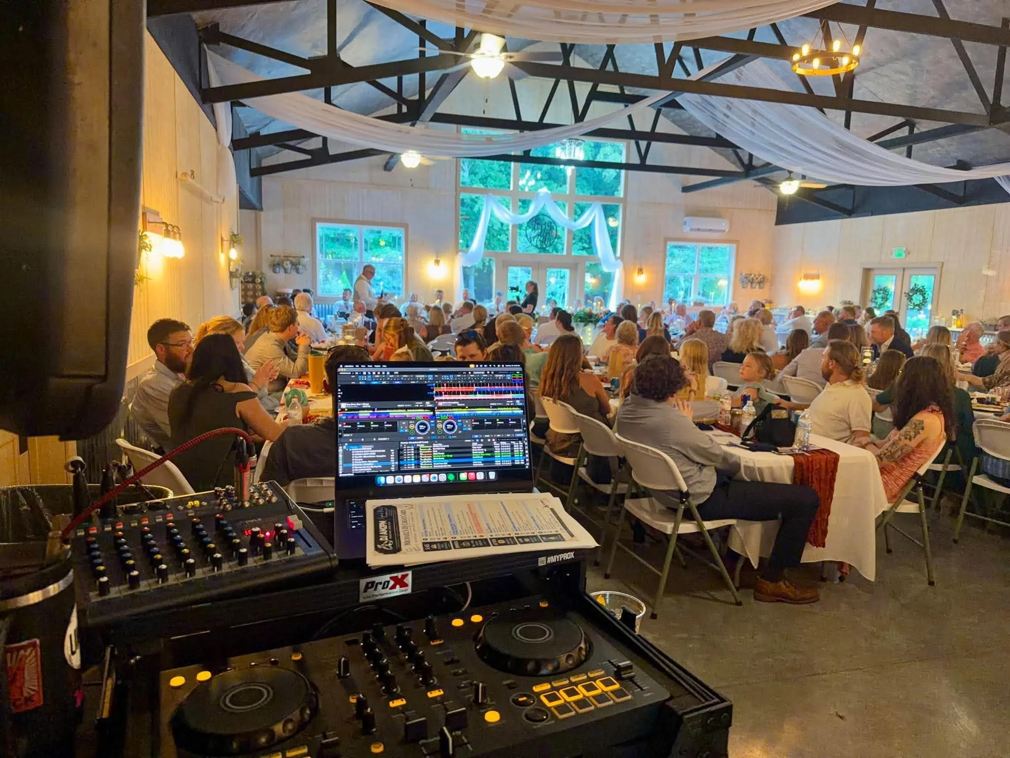 A large gathering of people seated at round tables in a decorated hall with draped fabric and large windows. In the foreground, DJ equipment and a laptop are visible, indicating a DJ setup for the event.