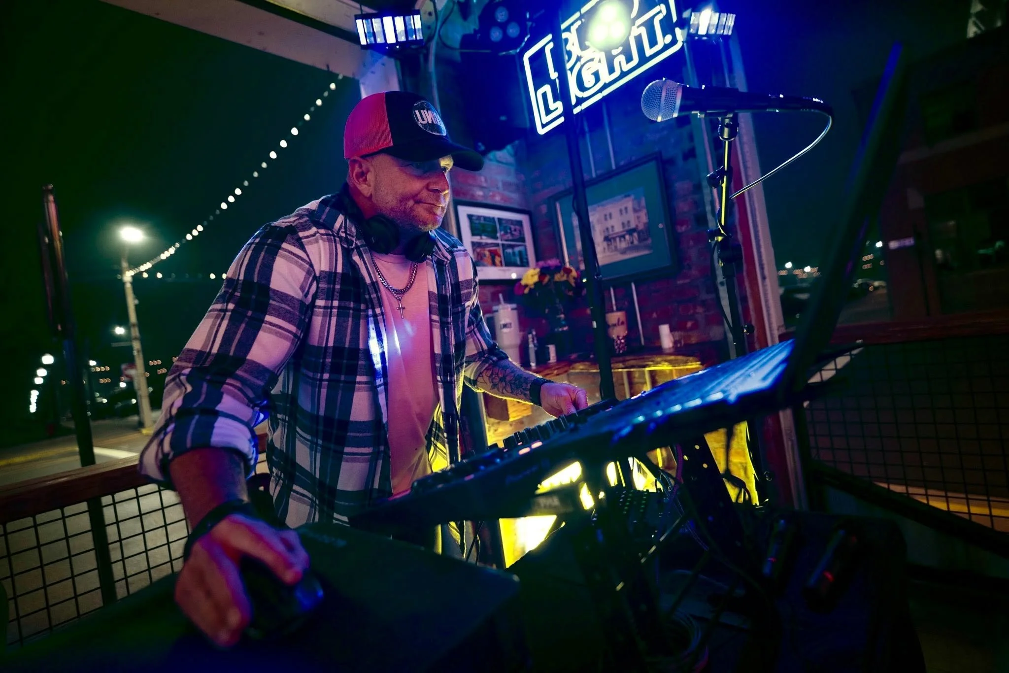 A DJ wearing a red and black trucker hat, plaid shirt, and headphones around his neck, is performing at a night event with a neon sign behind him that says 'Club'. He is in front of DJ equipment and a microphone, with city lights in the background.