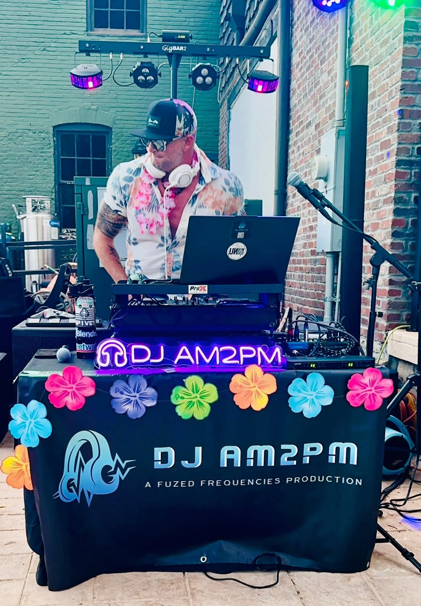 A DJ playing music at an outdoor event, with DJ equipment, a colorful neon sign that reads 'DJ AM3PM', and decorations of paper flowers on the table.