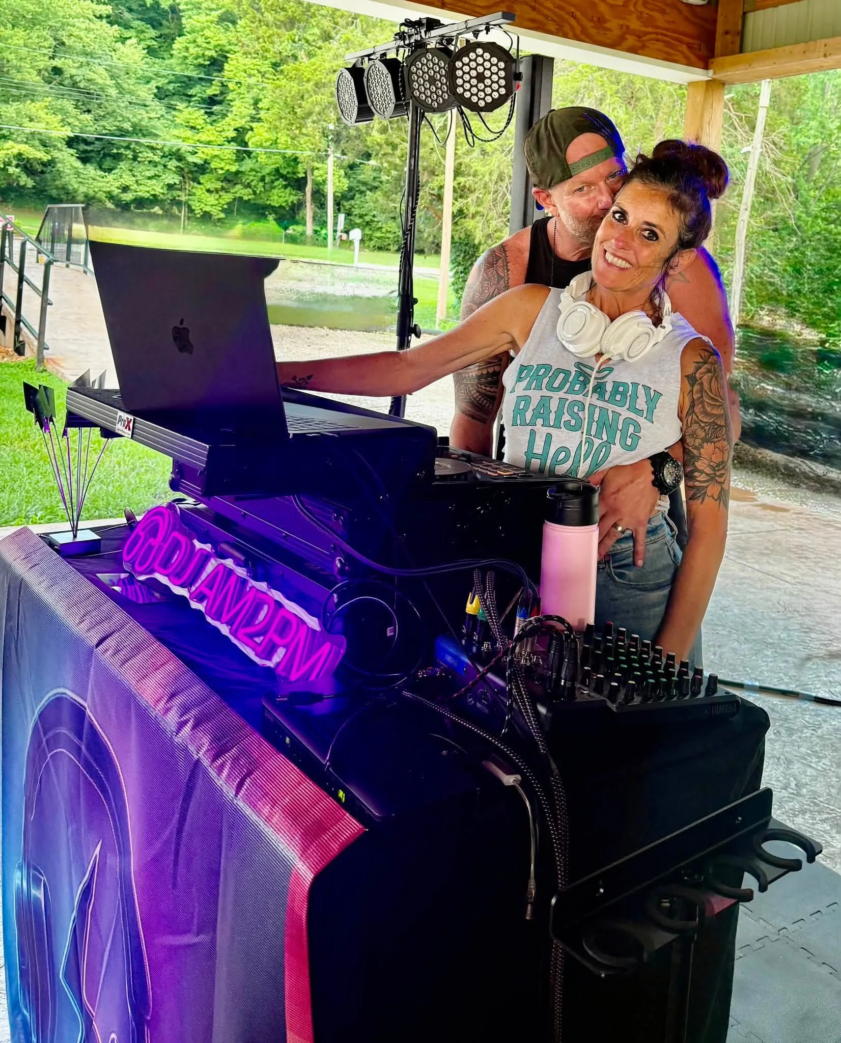 A woman smiling with headphones around her neck and a man standing behind her, playing a keyboard during an outdoor event on a covered porch, with trees and greenery in the background.