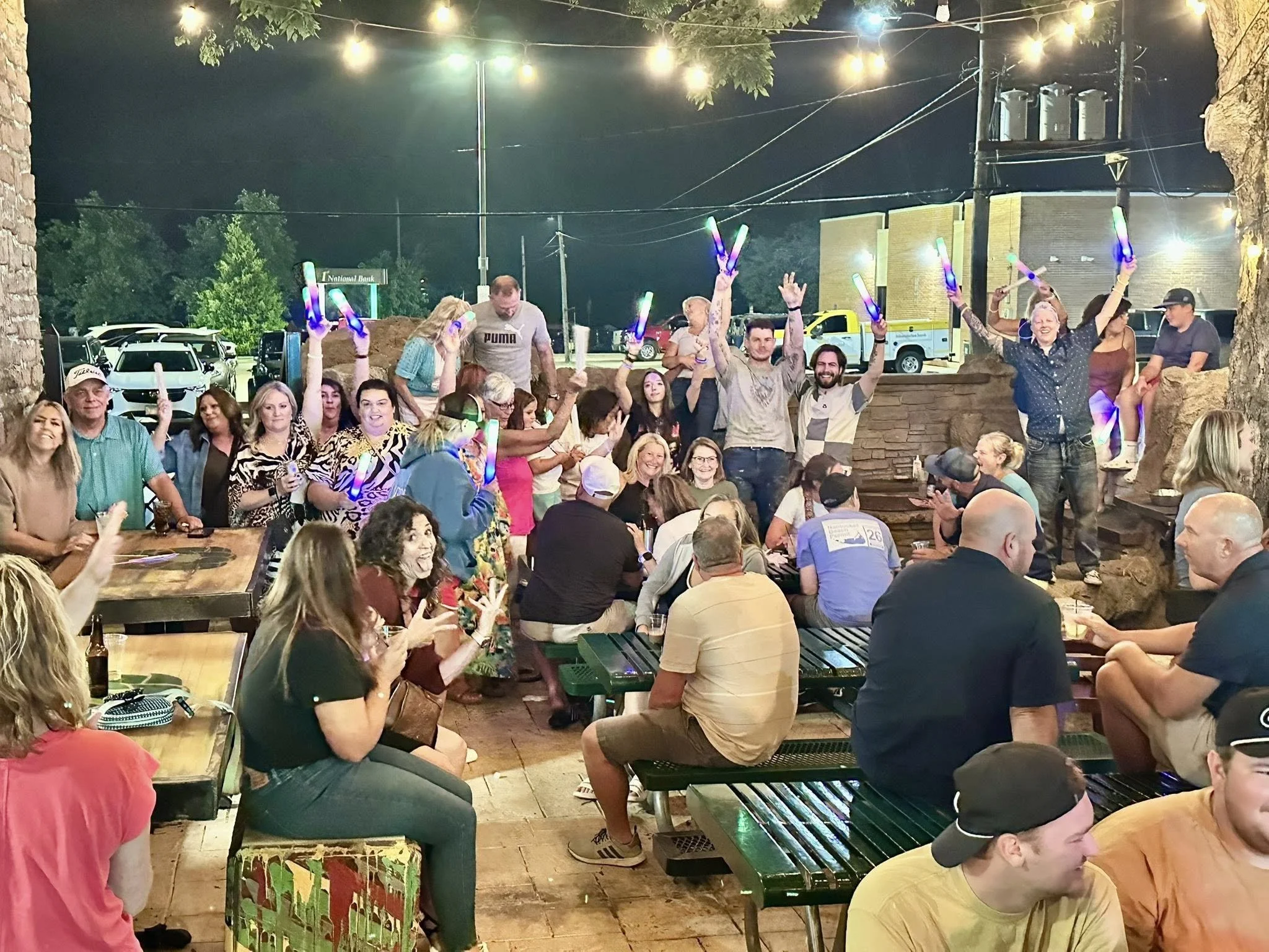 A large group of people gathered outdoors at night, some holding glow sticks and sitting at picnic tables, with string lights overhead, trees, parked cars, and a building in the background.
