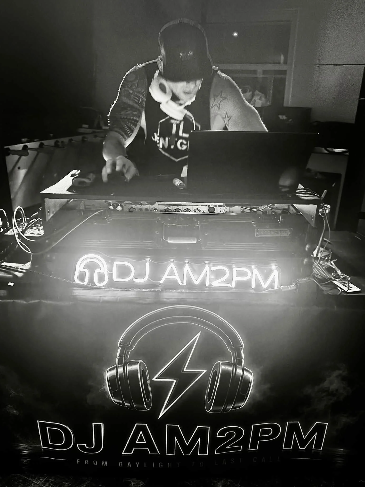 Black and white photo of a person wearing headphones, working with DJ equipment, with flashing neon-style text below that reads 'DJ AM2PM'. The background features a window and some furniture, creating a nightclub or studio vibe.