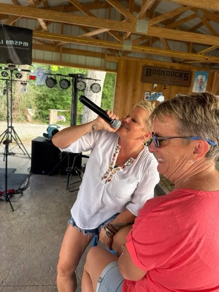Women singing and laughing at an outdoor covered venue with musical equipment and a sign that says 'BOONDOCKS'