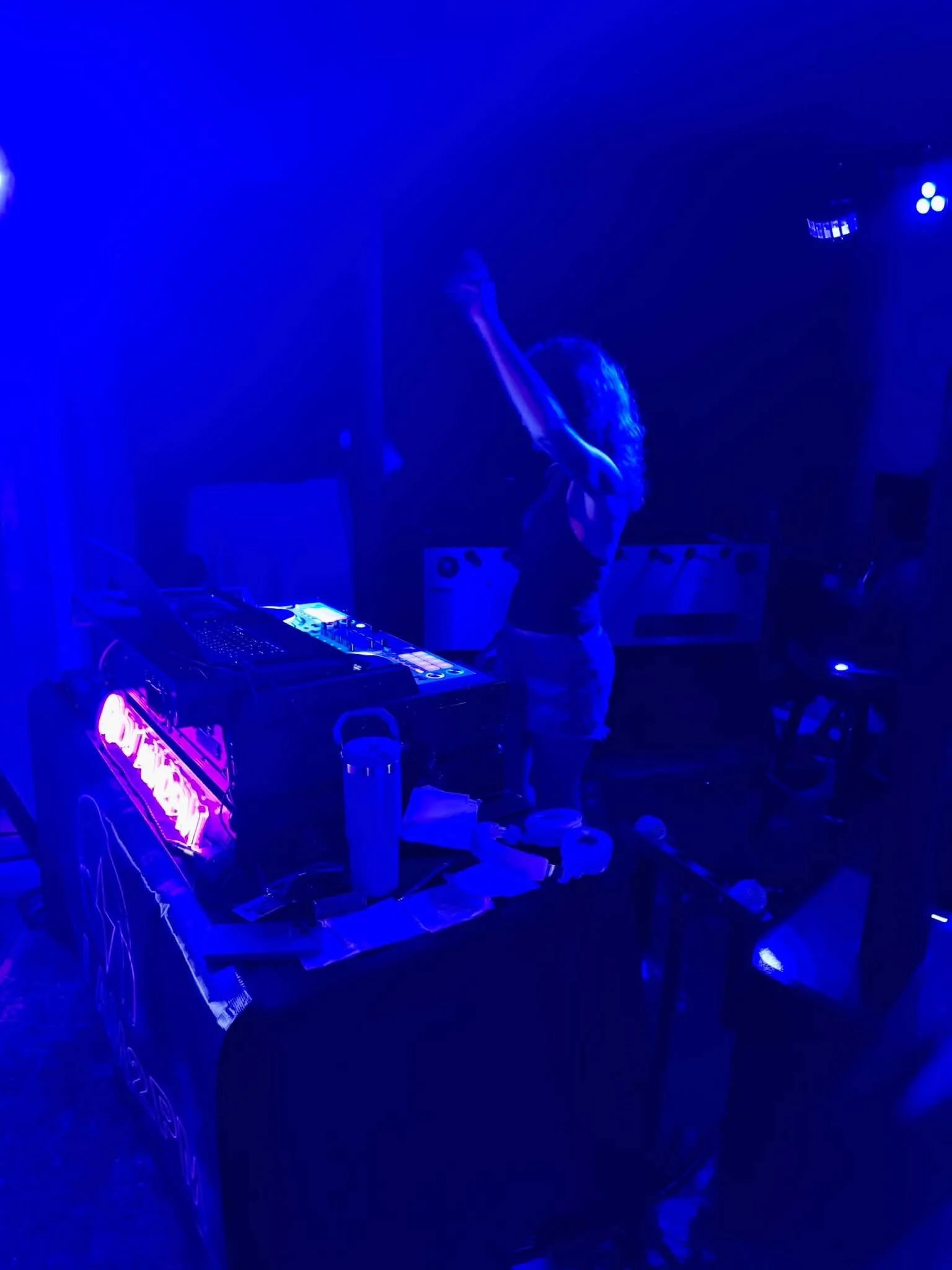 A woman singing into a microphone on a stage with blue lighting, a DJ setup, and various drinks on the table.